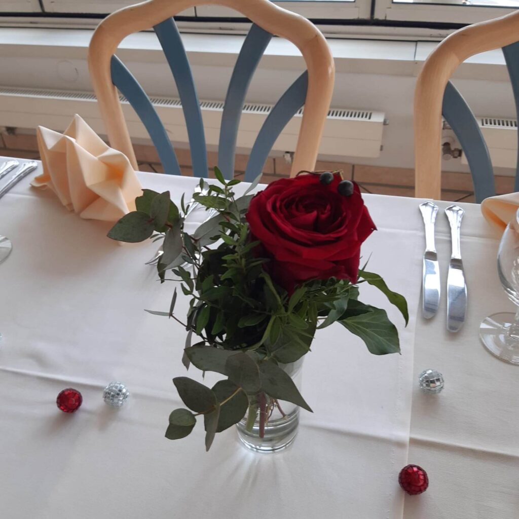 Bild einer Rose, diese steht auf einem Tisch in einer Vase, die Rose ist Rot.